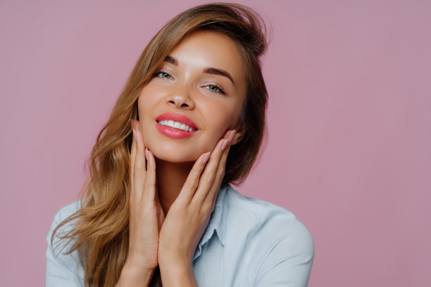 照片中可爱的年轻女模特双手轻触下巴，微笑温柔，面容柔美，长发梳在右侧，画着专业的妆容，紫色背景下孤立图片下载