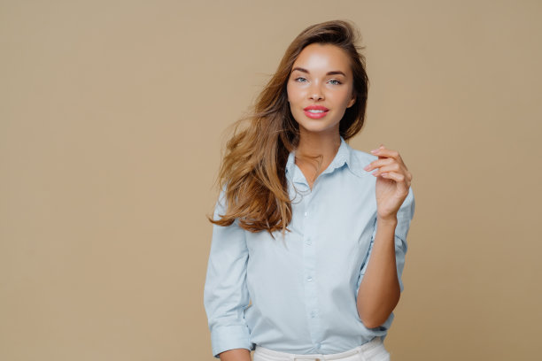 可爱的长发女子，手举着，穿着时尚的衬衫，平静的表情，微笑着，站在棕色背景的工作室里。人与时尚理念图片下载