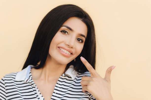近距离的肖像，高兴的黑发女性黑眼睛，漂亮的化妆，表明在嘴，有宽阔的微笑，示范白牙与牙套，显示她的美丽，孤立在空白的墙上图片下载