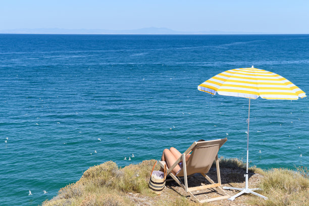 戴着草帽的女人坐在日光浴床上，躺椅上，在山顶的黄色伞下，悬崖上有蓝色的大海，海景。图片下载
