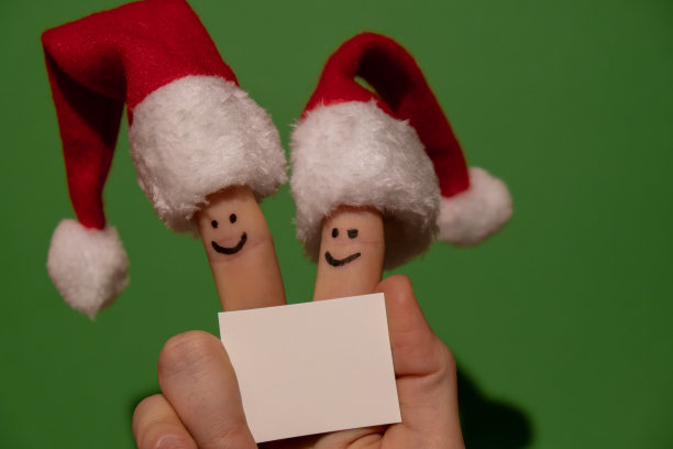 手指戴上圣诞老人的红白帽子。两个快乐的手指笑脸拿着空白的纸笔记为您的文本复制空间对绿色背景。圣诞节或新年的快乐家庭庆祝概念。夫妇图片下载