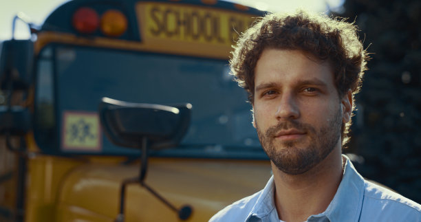 自信的男子司机站在黄色的公共汽车看着相机在阳光下近距离。图片下载