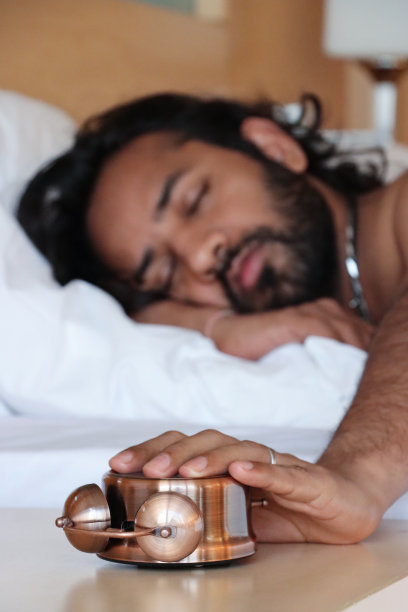 特写图像的印度男子躺在酒店的双人床下被褥，手伸向床头柜关闭打翻了青铜金属盒复古闹钟，双钟躺在脸朝下，焦点在前景图片下载