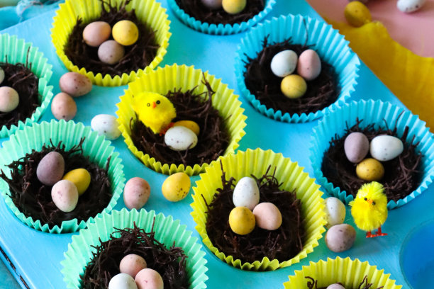 绿松石的特写图像，松饼烤盘上有一排排自制巧克力燕窝蛋糕，放在色彩鲜艳的纸蛋糕盒里，顶部有迷你巧克力复活节彩蛋，蓬松的黄色小鸡玩具装饰，黄色棉布，粉红色背景图片下载