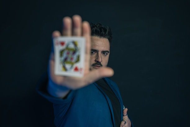 模糊的肖像，一个年轻的魔术师在黑暗中显示在他的手掌隐藏的卡，同时看着相机。他的脸很清晰。图片下载