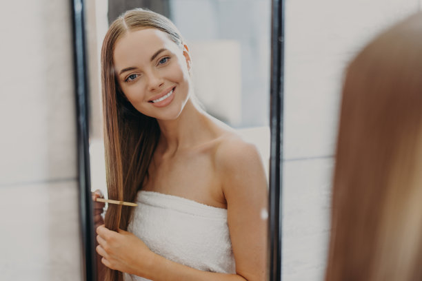 快乐美丽的年轻女子看着镜子里自己的倒影，有一头精心打理的长发，用梳子，裹着浴巾，做发型。女性，美容，美发和自我护理的概念图片下载