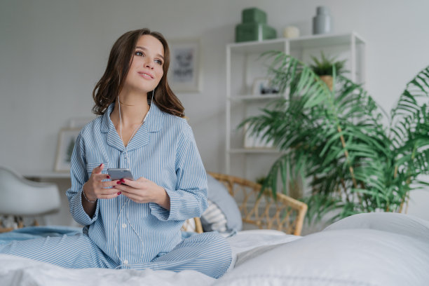 照片中，漂亮的黑发学生通过手机应用程序学习语言，戴着耳机听音频，有空闲时间，穿着休闲服装，躺在舒适的白色床上，目光深沉图片下载