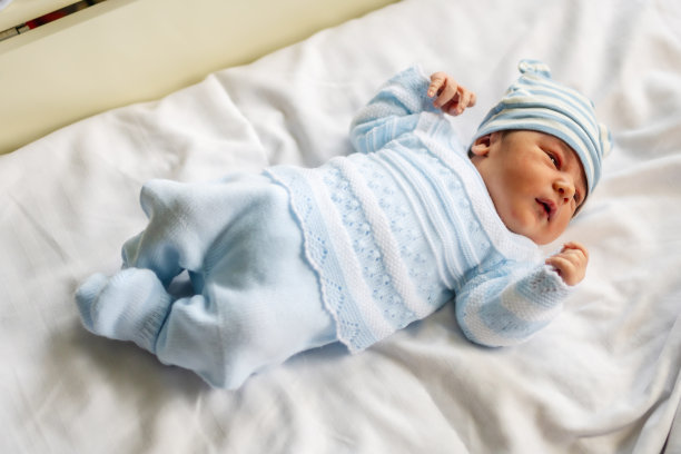医院里一个新生男孩的照片，他穿着白色的紧身衣，戴着蓝色的帽子，躺在婴儿床上图片下载
