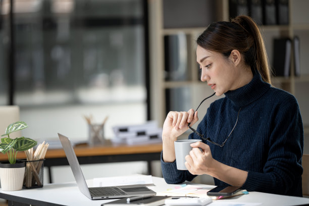 年轻的女商人在便装拿着杯子和眼镜，思考她的工作时，在办公室的笔记本电脑工作。图片下载