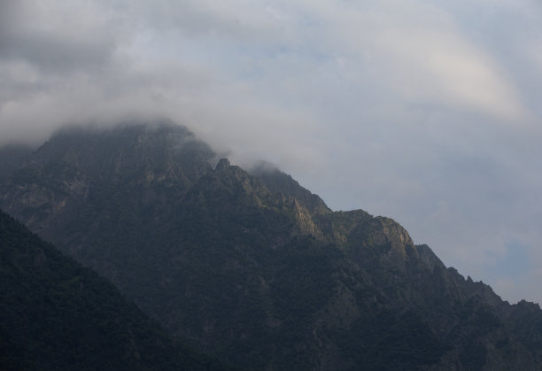 黑暗的大气超现实景观，在灰色多云的天空中，一个黑暗的岩石山峰在低云中。图片下载
