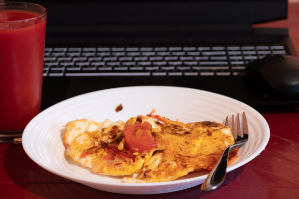 炒鸡蛋配一杯番茄汁放在红桌子上，他们拿了一台黑色笔记本电脑在家里的厨房里，一边工作一边有一台笔记本电脑图片下载