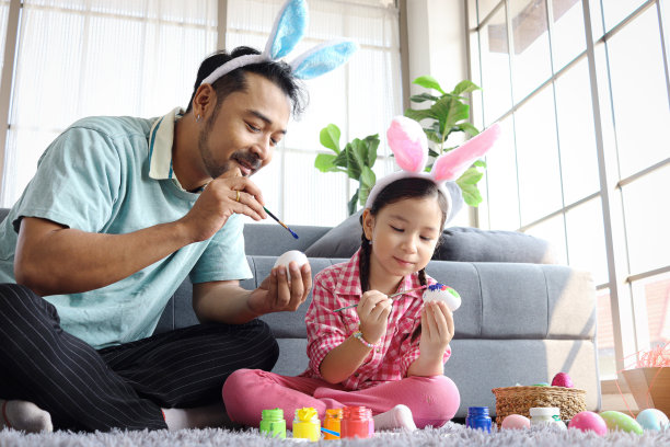 爸爸和女儿准备漂亮的复活节彩蛋装饰春天的庆典，粉红色的小兔子女孩和爸爸一起用兔子耳朵发带画彩色的彩蛋，快乐的复活节家庭图片下载