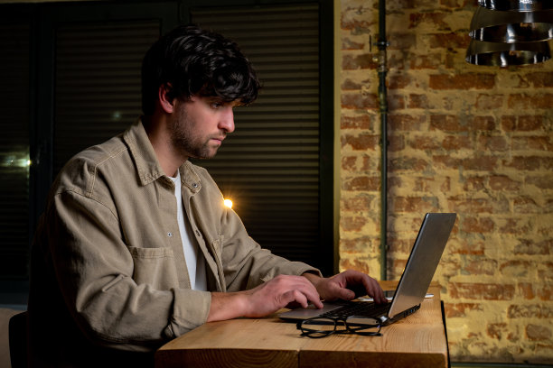 专注的男人在家里使用笔记本电脑。侧视图，一个穿着便服的男人坐在靠窗的桌子旁，在键盘上打字图片下载