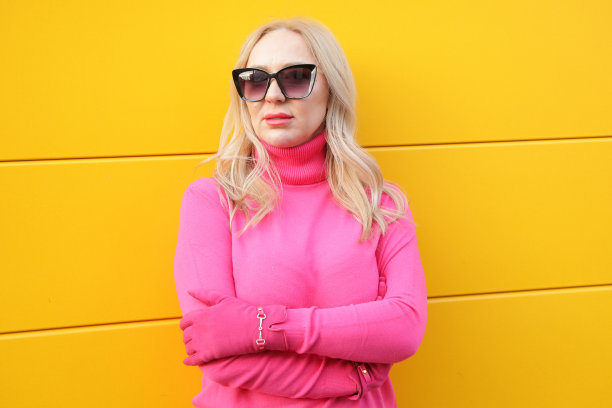 成功的金发女人在亮粉色高领毛衣和手套太阳镜在黄色的墙壁。色彩缤纷的休闲装。图片下载