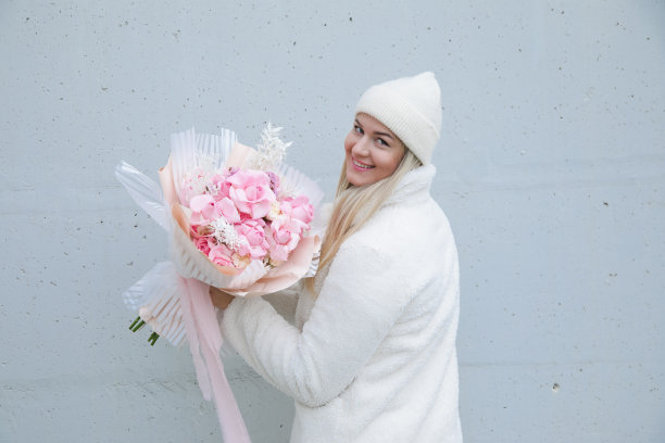 微笑的女人站在后面，手里拿着一束粉红色的花。灰色墙上的女性肖像。图片下载