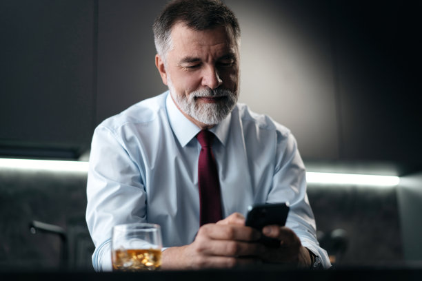 成熟的商人拿着一杯威士忌，坐在厨房里用智能手机。图片下载