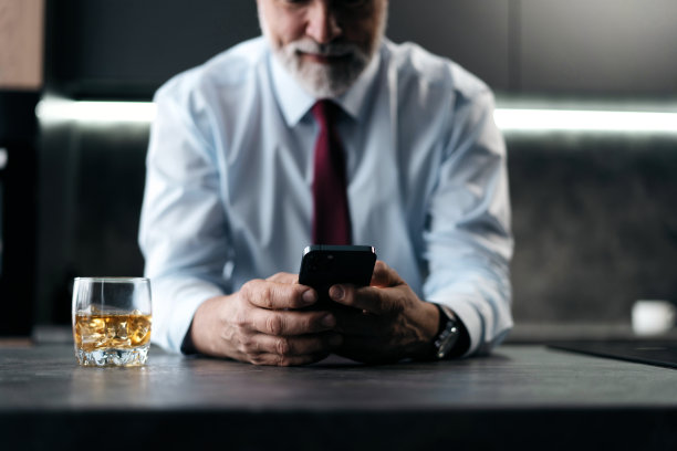 成熟的商人拿着一杯威士忌，坐在厨房里用智能手机。图片下载
