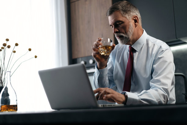 老男人一边喝威士忌，一边在厨房用笔记本电脑工作。图片下载