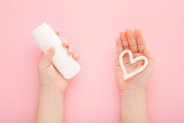 在浅粉色的桌子背景上，白色心形的女婴手掌。柔和的颜色。手持管保湿霜。特写镜头。视角镜头。关爱孩子双手肌肤。自顶向下视图。图片下载