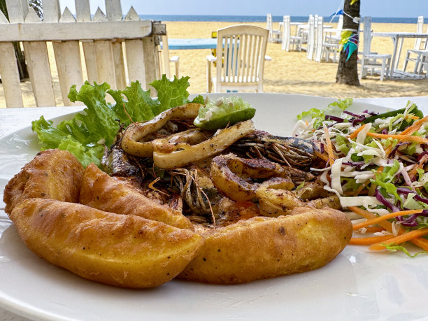 海滩餐厅酒吧场景的特写图像，俯瞰大海，沙滩和躺椅，金棕色，新鲜脆脆的薯条/薯条，炸鱿鱼圈和大虾放在白色盘子里，配上凉拌卷心菜和生菜，青柠角，重点在前景图片下载