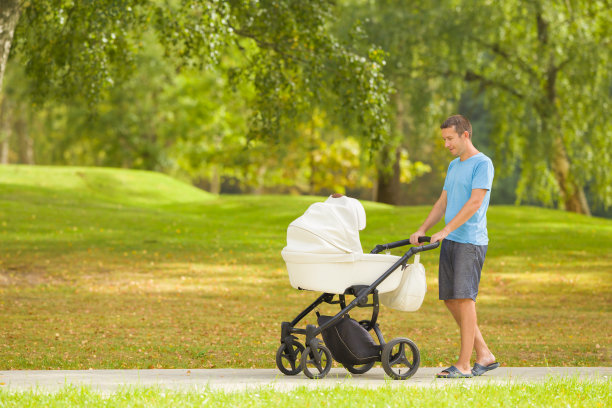 年轻的成年父亲推着白色的婴儿车，慢慢走在小镇公园的人行道上，在温暖阳光明媚的夏日。陪伴新生儿，呼吸新鲜空气。享受漫步。侧视图。图片下载