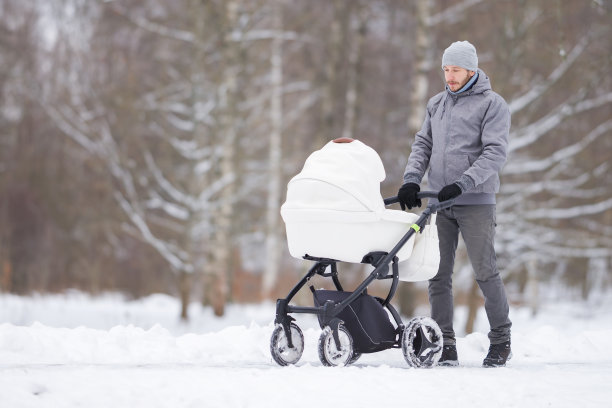 微笑的年轻成年父亲推着白色的婴儿车，在寒冷的冬天走在公园积雪覆盖的人行道上。陪伴新生儿，呼吸新鲜空气。享受宁静的散步。图片下载