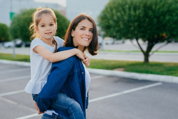 在户外拍摄的照片中，微笑的女人背着女儿，在户外散步，积极地微笑，一起玩耍和玩得很开心，在沥青和绿色的树木背景下摆姿势。家庭的概念图片下载