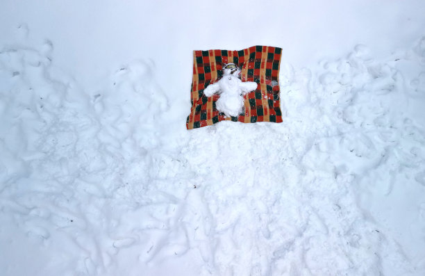 一个雪人躺在毯子上，双臂伸开，正在晒太阳。他有太阳镜和雪茄。在海滩或冬季滑雪胜地度假。从山上用无人机飞行。图片下载