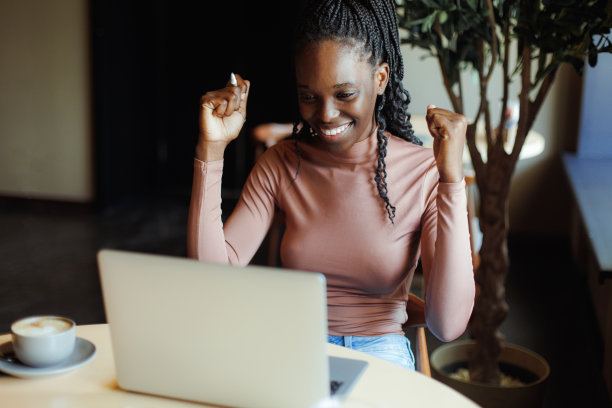 快乐的非洲裔美国妇女高兴弯曲肘部，坐在桌子上，在舒适的咖啡馆里对着笔记本电脑说话。黑人女孩远程在线工作，自由职业者。幸运相逢，视频通话，成功图片下载