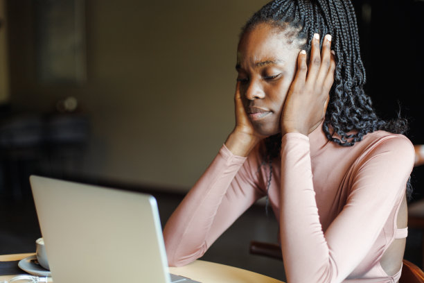 年轻疲惫的非裔美国妇女坐在桌旁，看着笔记本电脑的屏幕，手放在脸颊上。图片下载