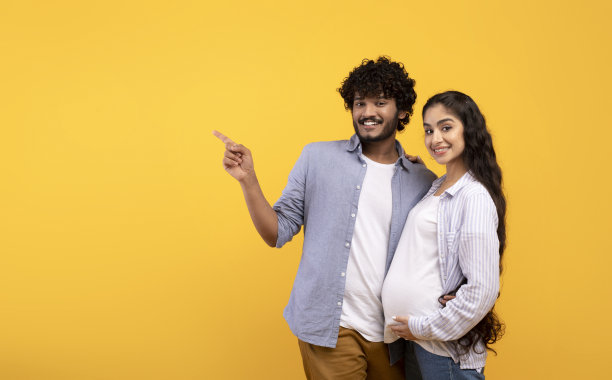 年轻可爱的印度怀孕夫妇指着复印空间，显示伟大的优惠或促销，黄色背景图片下载
