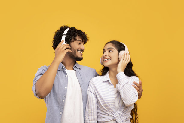 一对可爱的印度夫妇戴着耳机听音乐，在黄色的演播室背景下拥抱、看着对方图片下载