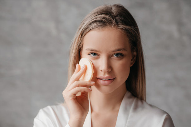 护肤概念。年轻女子使用化妆海绵，同时做面部护肤程序，站在浴室图片下载