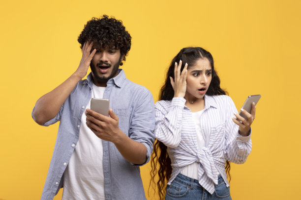 一名印度女子和她的男友震惊地看着他们的智能手机，在黄色背景下触摸头部图片下载