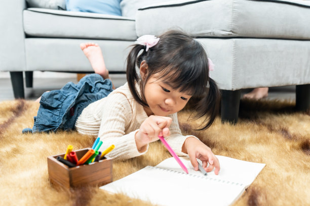 可爱的亚洲女孩躺在房子地板上的地毯上，手里拿着彩色铅笔，快乐地在房子客厅的笔记本上画画。家庭幸福图片下载