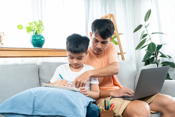 一名亚洲男子正在家里客厅的沙发上引导他的儿子做作业。当爸爸在家用他的笔记本电脑上网工作的时候，单身父亲在指导你的儿子。图片下载