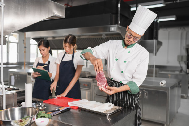 肖像亚洲年轻男子在厨师制服烹饪牛排火与学生女助理厨师在厨房图片下载