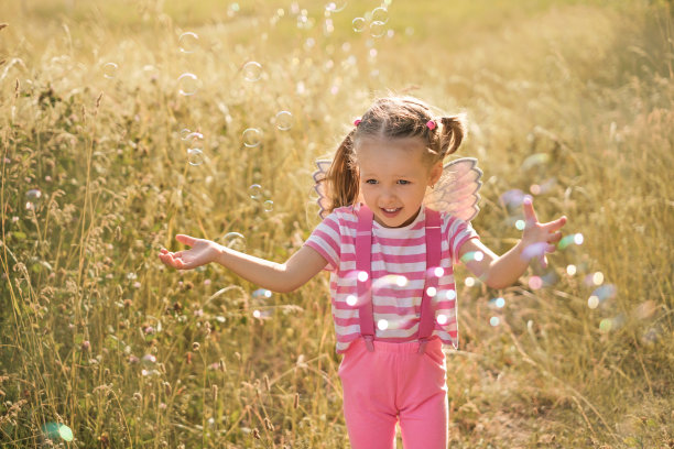背上长着翅膀的有趣女孩跑过草地，抓肥皂泡图片下载