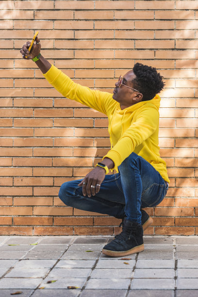 年轻黑人对着砖墙用智能手机自拍图片下载