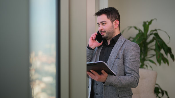 年轻的商人站在窗户旁边，一边打电话一边检查文件和现代办公室图片下载
