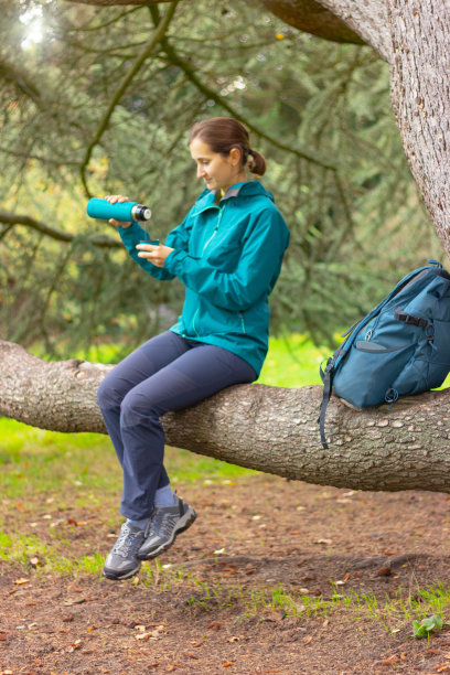 一名年轻女子坐在森林里的树枝上，用保温杯把热茶倒进杯子里，人的视线模糊了。图片下载