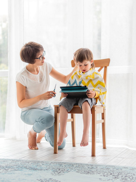 母亲亲自给儿子理发。小男孩拿着平板电脑坐着。新常态图片下载