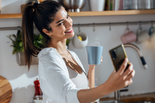 美女一边在家里的厨房里喝咖啡，一边用手机自拍。图片下载