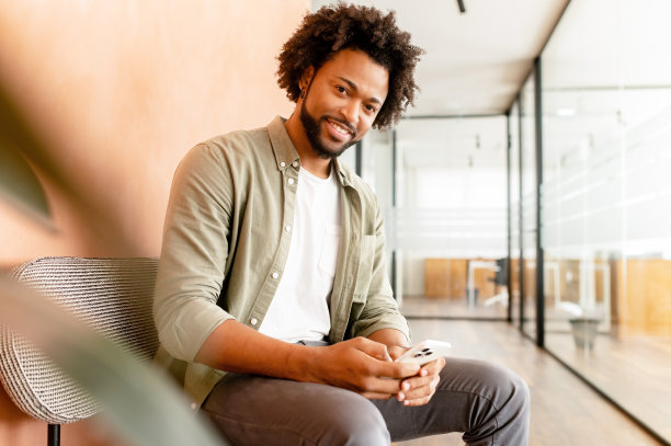 微笑的非洲裔美国人坐在办公室的沙发上，用手机检查社交网络，为家人，女朋友，商业伙伴输入信息，查看照片图片下载