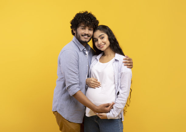 满意的年轻印度男子拥抱怀孕的妻子，摸着肚子，在黄色背景下对着摄像机微笑图片下载
