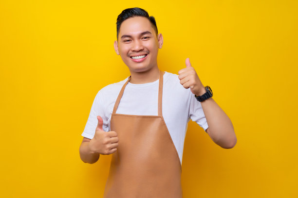穿着棕色围裙的年轻亚裔男子在咖啡店工作，看着镜头，在黄色背景下竖起大拇指表示赞同和推荐。小型企业创业概念图片下载