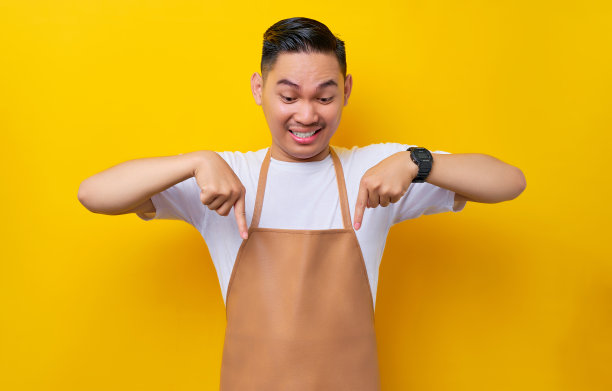 快乐的年轻亚洲男子20多岁，穿着棕色围裙的咖啡服务员在咖啡店工作，手指指向黄色背景的工作区。小型企业创业概念图片下载