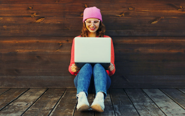 时尚的现代年轻女子的肖像与笔记本电脑穿着彩色的衣服坐在城市公园的木墙背景图片下载