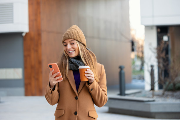 年轻女子与智能手机站在街道模糊的建筑背景。漂亮女孩的时尚商业照片与手机和一杯咖啡图片下载