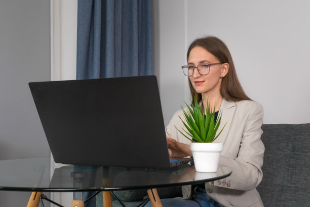 年轻迷人的女人戴着眼镜，一件夹克坐在家里的桌子上，在一台笔记本电脑前工作。理念学习，远程办公图片下载